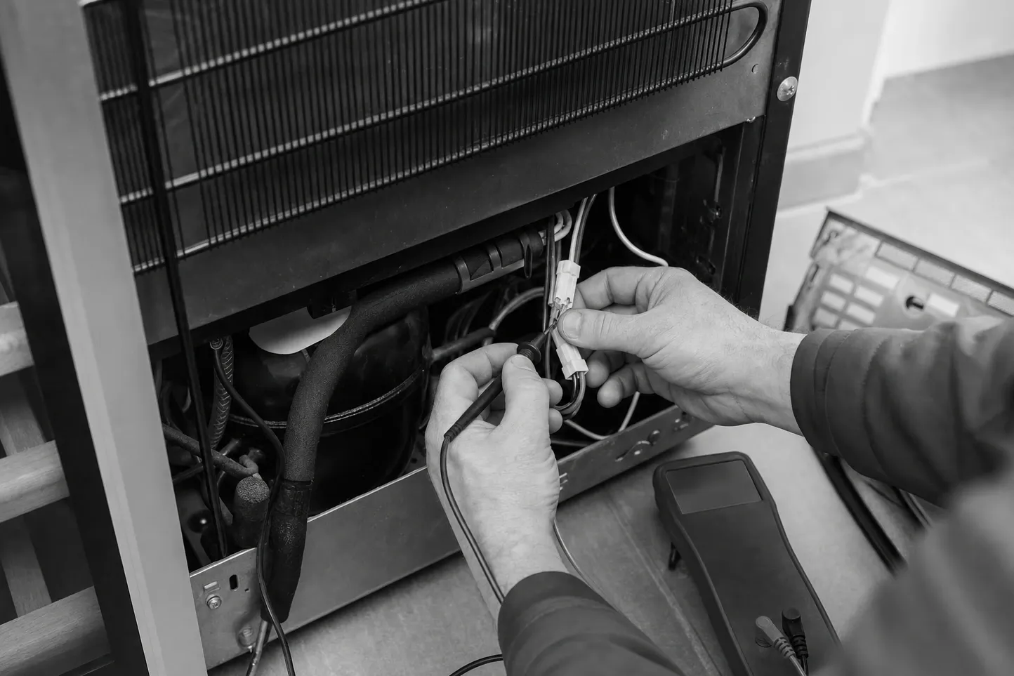 Black-and-white close-up of both hands separating a wiring connector at the rear machine compartment of a refrigerator, condenser coil above