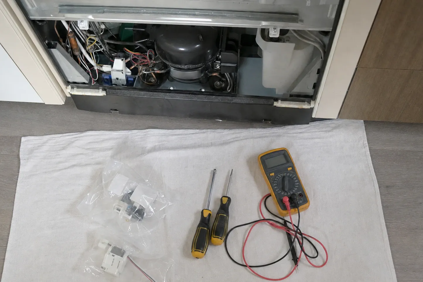 Open lower machine compartment seen from above, with two bagged replacement parts, screwdrivers and a multimeter laid out on a drop cloth