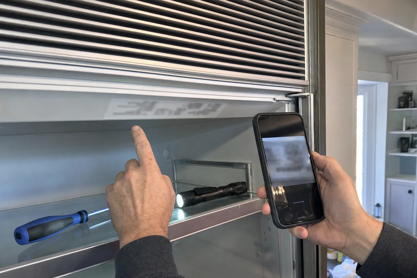 One hand pointing at the rating plate inside the top of an open built-in refrigerator while the other photographs it with a phone
